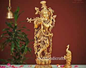 May include: A gold-colored metal statue of Krishna playing a flute, standing on a decorative base. The statue is intricately detailed and features a peacock design. The statue is surrounded by pink rose petals.