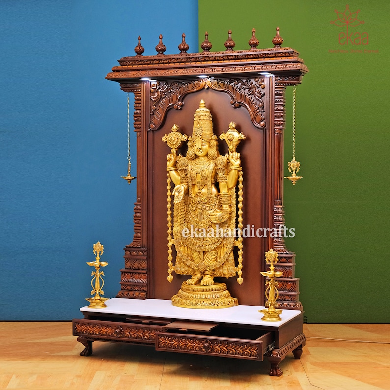 May include: A wooden shrine with intricate carvings and a gold-colored statue of a deity. The shrine has a white marble top and a drawer. The statue is holding two objects in its hands.