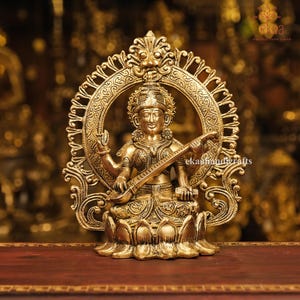 Può includere: Una statua in ottone dorato di una divinità, seduta su una base a forma di fiore di loto, che tiene uno strumento a corde. La statua è incorniciata da un'aureola circolare ornata. L'immagine ha il testo "ekahandicrafts".