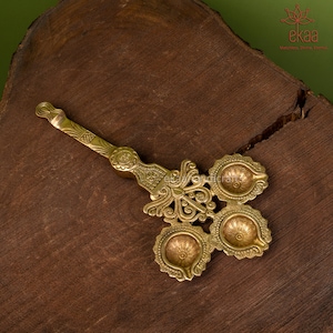 May include: A gold-colored metal diya with four wicks, each with a decorative floral design. The diya has a long handle with intricate details.