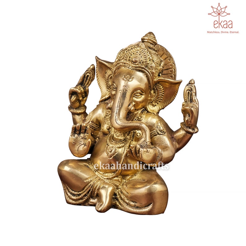 May include: A gold-colored brass statue of Ganesha, the Hindu god of wisdom and prosperity, sitting in a meditative pose with his trunk raised and his hands in a gesture of blessing. The statue is intricately detailed with decorative patterns and a crown.