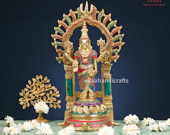 May include: A gold-colored statue of a Hindu deity with a red and green patterned dress, standing on a base with a red and green mosaic design. The statue is surrounded by a decorative archway with red and green accents. The statue is on a wooden table with white flowers.