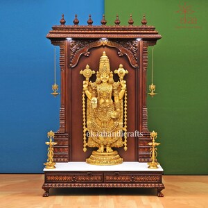 May include: A wooden temple shrine with intricate carvings and a gold-colored deity statue inside. The shrine has two drawers and a marble top. The deity is holding a staff in each hand and is adorned with jewelry.