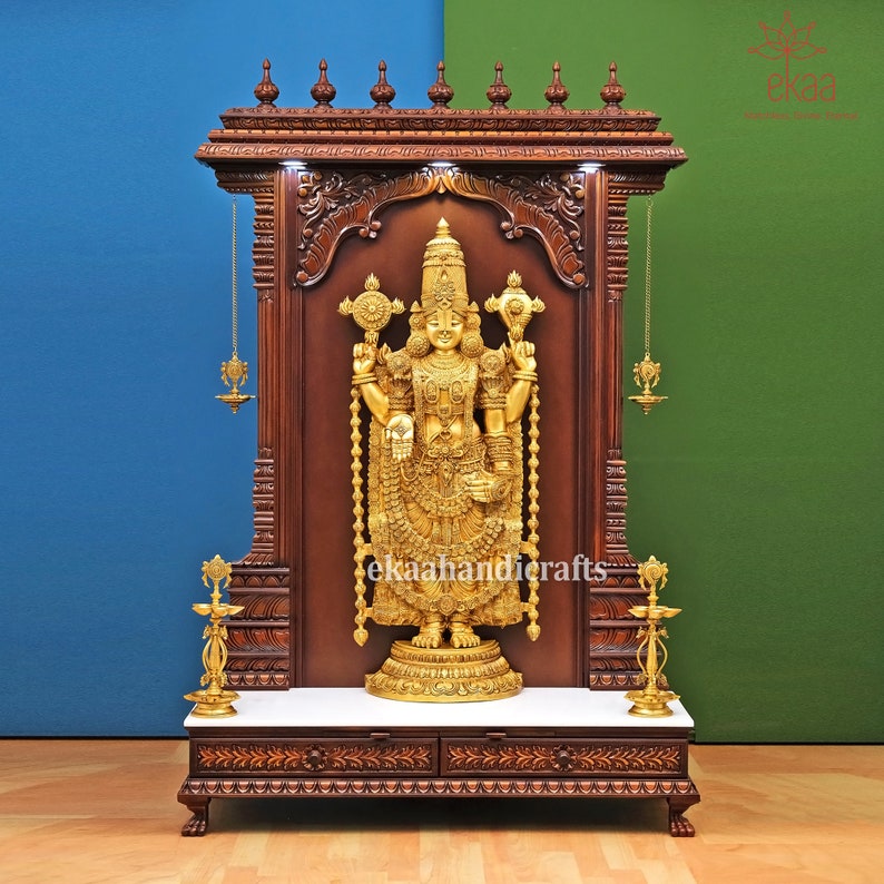 May include: A wooden temple shrine with intricate carvings and a gold-colored deity statue inside. The shrine has two drawers and a marble top. The deity is holding a staff in each hand and is adorned with jewelry.