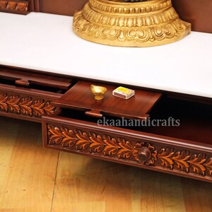 May include: A wooden altar with a white marble top and a drawer with intricate carvings. The drawer is open and contains a small golden bowl and a box of matches. The text "ekaahandicrafts" is visible on the drawer.