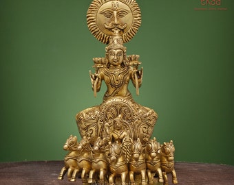 May include: A gold-colored metal statue of the Hindu deity Surya, the Sun God, riding a chariot pulled by seven horses. Surya is depicted with a crown and a halo, and he is holding a lotus flower in each hand. The statue is on a wooden surface.