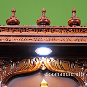 May include: A wooden shrine with intricate carvings and a white light shining from the top. The shrine has three decorative finials and the text "ekaahandicrafts" is visible at the bottom.