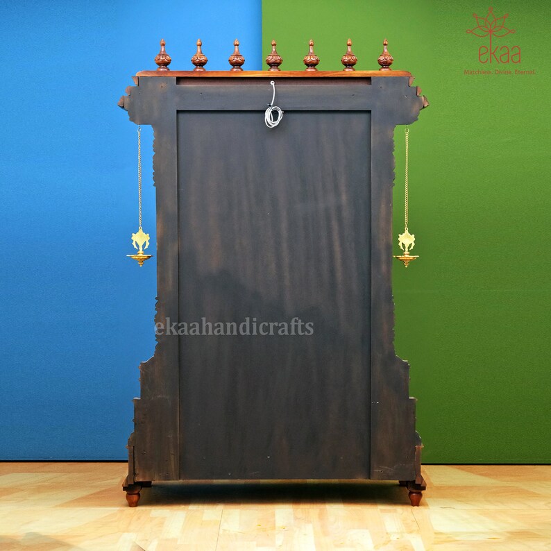 May include: A dark wood temple-style cabinet with intricate carvings and gold accents. The cabinet has two doors and a shelf inside. The cabinet is decorated with two brass oil lamps hanging from chains.