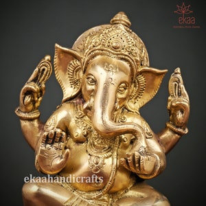 May include: A gold-colored brass statue of Ganesha, the Hindu god of beginnings, wisdom, and prosperity. The statue depicts Ganesha sitting with his trunk raised and his hands in a mudra, or symbolic gesture. The statue is intricately detailed with floral patterns and other decorative elements. The statue is made by ekaahandicrafts.