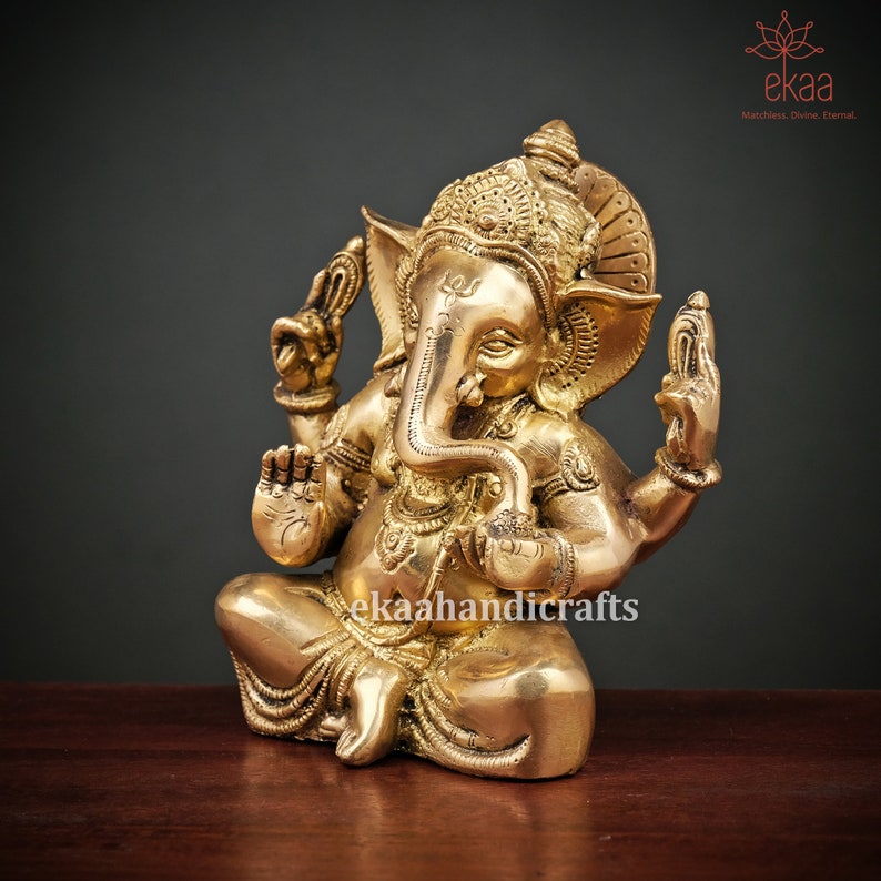 May include: A gold-colored brass statue of Ganesha, the Hindu god of wisdom and prosperity, sitting in a meditative pose with his trunk curled up. He is adorned with intricate details and a crown.