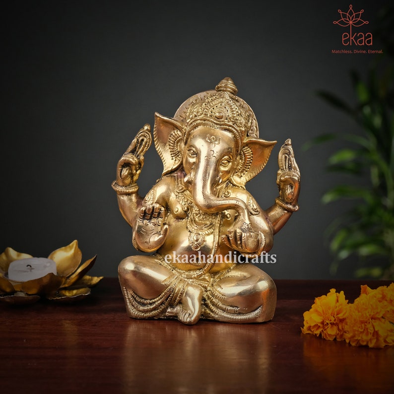 May include: A gold-colored brass statue of Ganesha, the Hindu god of wisdom and prosperity, sitting in a meditative pose with his trunk raised. The statue is intricately detailed with floral patterns and has a small, round base.