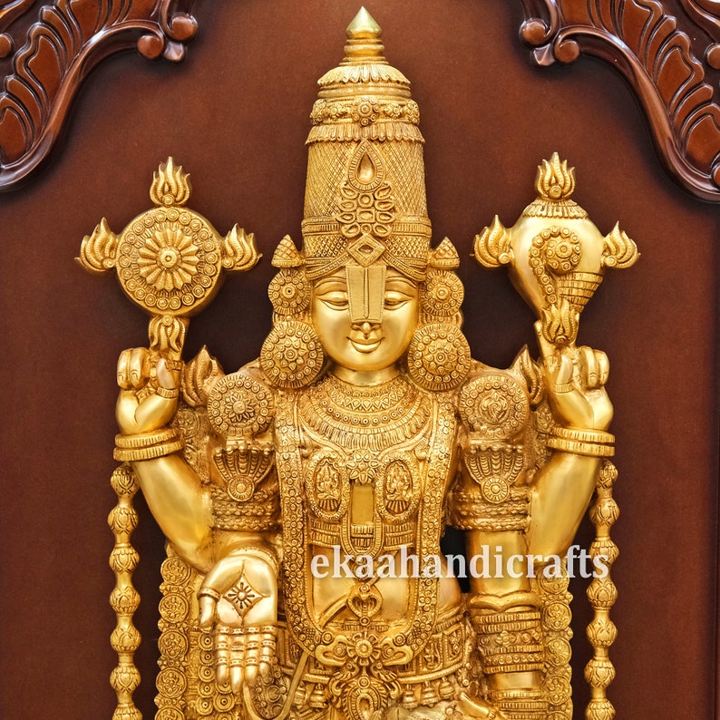 May include: A gold-colored metal sculpture of a Hindu deity, Vishnu, standing with his arms raised, holding a conch shell and a discus. The sculpture is intricately detailed and has a traditional Indian design. The sculpture is mounted on a wooden frame.