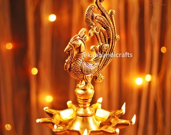May include: A gold metal diya with a peacock design. The diya has seven wicks and is lit with flames. The diya is a traditional Indian lamp used for religious ceremonies and festivals.
