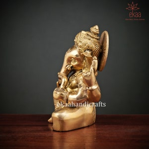 May include: A gold-colored brass statue of Ganesha, the Hindu god of wisdom and prosperity, sitting with his trunk raised and holding a sweet in his hand. The statue is intricately detailed and has a traditional Indian design.
