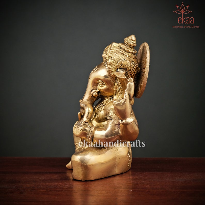 May include: A gold-colored brass statue of Ganesha, the Hindu god of wisdom and prosperity, sitting with his trunk raised and holding a sweet in his hand. The statue is intricately detailed and has a traditional Indian design.