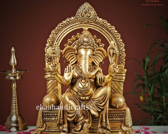 May include: A gold-colored brass statue of Ganesha, the Hindu god of wisdom and prosperity, sitting on a throne with intricate carvings. The statue is set against a brown background with rose petals scattered in the foreground.