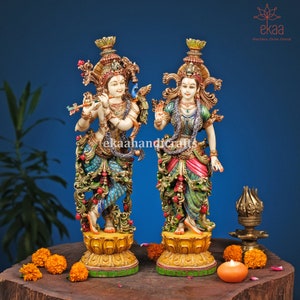 Puede incluir: Un par de estatuas de Radha y Krishna finamente talladas, de pie sobre bases ornamentadas. Las estatuas están pintadas en colores vibrantes, incluyendo oro, rojo, verde y azul. Las estatuas están hechas de madera y son un hermoso ejemplo de la artesanía india.