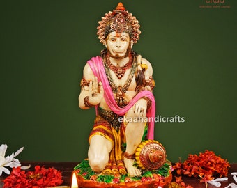 Może przedstawiać: Kolorowa statua hinduskiego bóstwa Hanuman, klęczącego z dłońmi w pozycji modlitewnej. Ma na sobie różowo-żółty dhoti i złoty naszyjnik. Statua stoi na brązowej podstawie z zielonymi liśćmi i kwiatami.