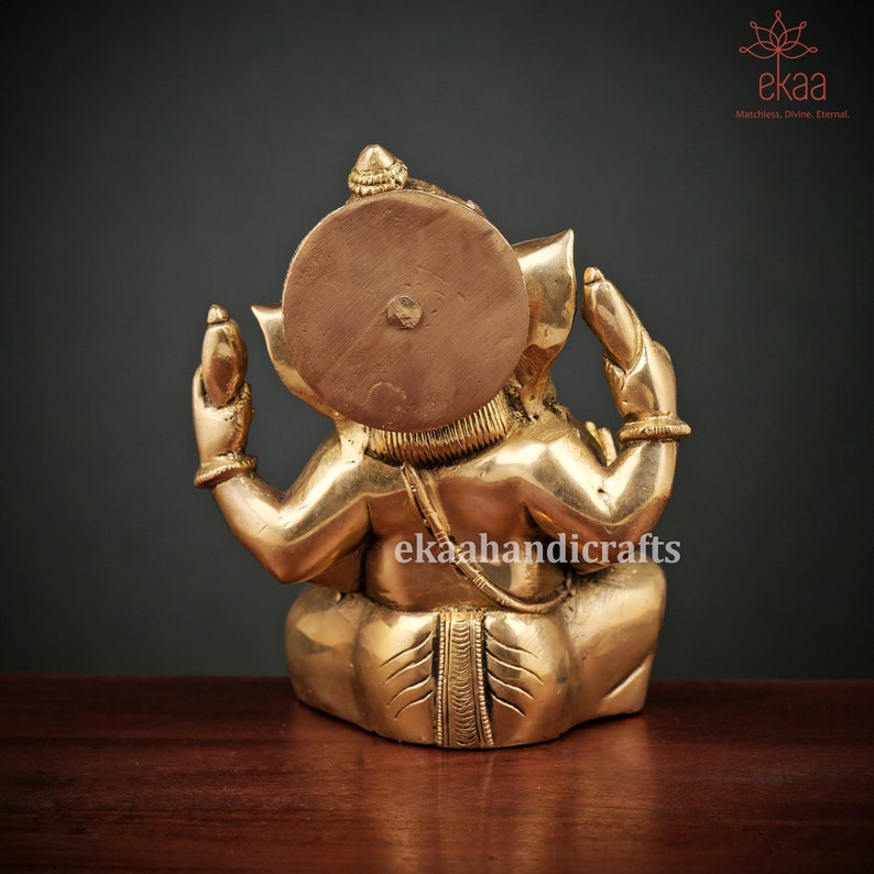 May include: A gold-colored brass statue of Ganesha, the Hindu god of beginnings, wisdom, and prosperity. The statue is sitting with its legs crossed and its hands in a mudra, a symbolic gesture. The statue is decorated with intricate details and has a round, flat disc behind its head.