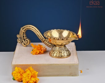 May include: A gold brass diya with a decorative handle, lit with a flame, sitting on a book with yellow flowers.