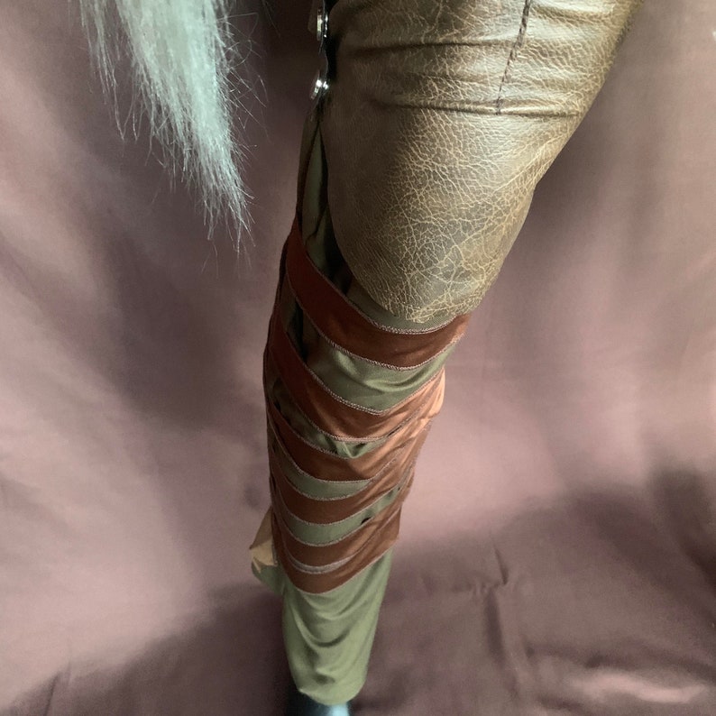 Puede incluir: Pantalones de cuero marr&oacute;n con un panel de tela verde y correas marrones. Las correas est&aacute;n superpuestas y se cruzan por la pierna.