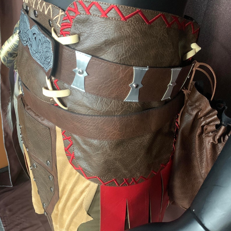 Puede incluir: Un cintur&oacute;n de cuero marr&oacute;n con detalles met&aacute;licos decorativos y costuras rojas. El cintur&oacute;n se lleva sobre una falda de cuero marr&oacute;n con un panel de tela beige y flecos rojos.
