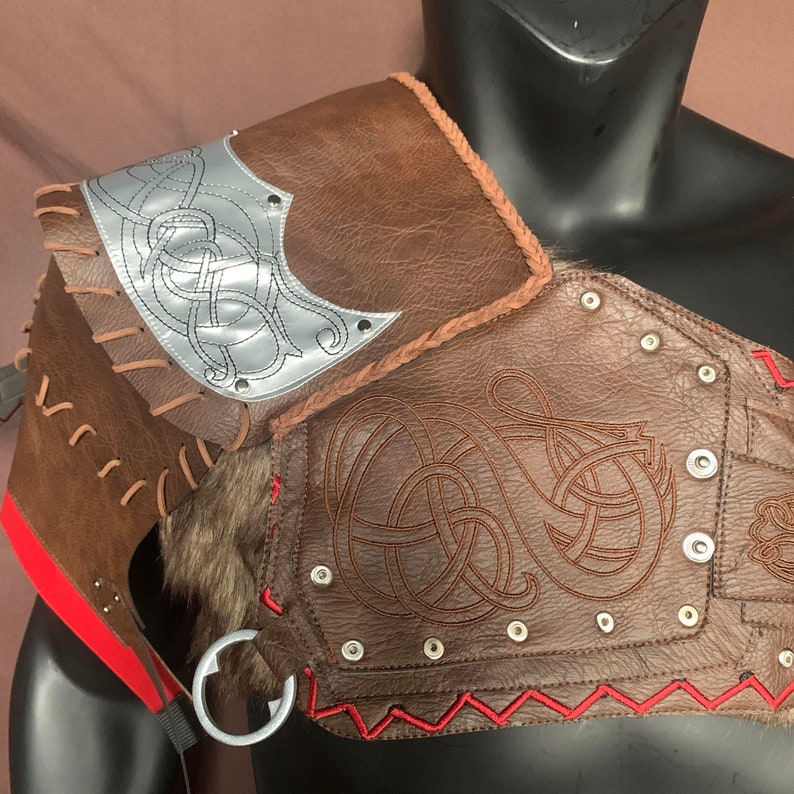 Puede incluir: Armadura de hombro de cuero marr&oacute;n con un dise&ntilde;o plateado y costuras rojas. La armadura tiene un dise&ntilde;o de nudo celta y est&aacute; decorada con remaches.
