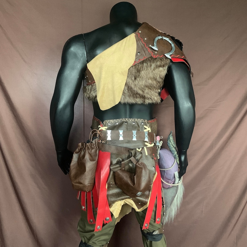 Puede incluir: Un traje de cuero marr&oacute;n con un chaleco de piel, un cintur&oacute;n con detalles met&aacute;licos y correas rojas. El traje lo lleva un maniqu&iacute;.