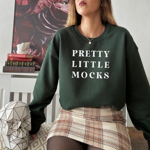 Op de afbeelding: Een donkergroen sweatshirt met de tekst "PRETTY LITTLE MOCKS" in wit gedrukt op de voorkant. Het sweatshirt wordt gedragen met een geruite rok.
