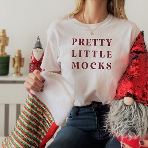 Puede incluir: Una camiseta blanca con el texto "PRETTY LITTLE MOCKS" impreso en granate. La persona lleva pantalones vaqueros azules y sostiene una media rayada roja y verde y un gnomo rojo con lentejuelas.