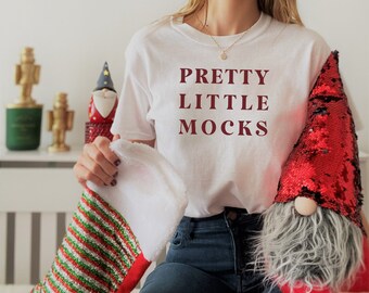 Könnte beinhalten: Ein weißes T-Shirt mit dem Text "PRETTY LITTLE MOCKS" in Rotbraun bedruckt. Die Person trägt eine blaue Jeans und hält einen rot-grünen gestreiften Strumpf und einen roten, mit Pailletten besetzten Zwerg.