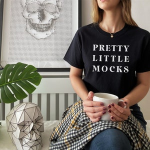 May include: A person wearing a black t-shirt with the text "Pretty Little Mocks" in white lettering. The person is holding a white ceramic mug and is sitting on a checkered blanket.