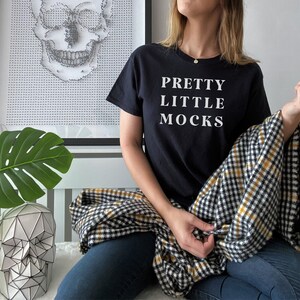May include: A black t-shirt with the text "PRETTY LITTLE MOCKS" printed in white on the front. The person wearing the shirt is sitting on a black and white checked blanket.