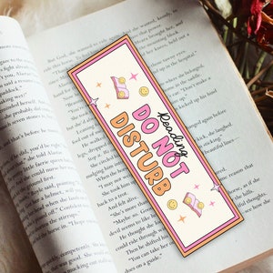 May include: A rectangular bookmark with a pink border and the words "DO NOT DISTURB" in orange and pink. The bookmark has a retro design with smiley faces, stars, and a book graphic. The words "Reading" are written above the main text.