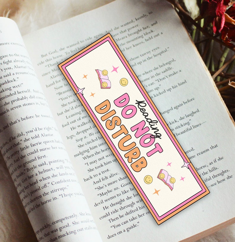 May include: A rectangular bookmark with a pink border and the words "DO NOT DISTURB" in orange and pink. The bookmark has a retro design with smiley faces, stars, and a book graphic. The words "Reading" are written above the main text.
