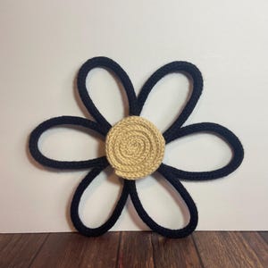 May include: A handcrafted, decorative flower with a navy blue rope design for the petals and a tan, coiled rope center. The flower is displayed against a white background and a wooden surface.