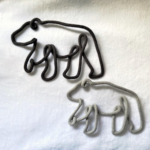 May include: Two bear-shaped wall hangings made from dark gray and light gray rope. The bears are outlined with the rope, creating a minimalist design. The background is a soft, white, textured surface.