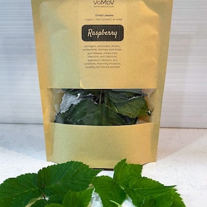 May include: A tan stand-up pouch labeled VoMoV containing dried raspberry leaves, with a clear window showing the contents. The label lists organic, hand-picked, and air-dried features. Several fresh green raspberry leaves are arranged in front.