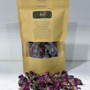 Op de afbeelding: Een bruine papieren zak met een doorzichtig venster, gevuld met gedroogde rozenknoppen. De zak is gelabeld met "Dried Flowers" en "Rose". Een hoop gedroogde, dieproze rozenknoppen ligt voor de zak. De tekst op de zak somt de voordelen van het product op.