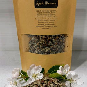 Op de afbeelding: Een kraftpapieren zakje met gedroogde appelbloesem met een doorzichtig venster, met het label "Apple Blossom". Een stapel gedroogde bloesem en verse witte appelbloesem liggen ervoor. De tekst op de zak vermeldt de toepassingen.