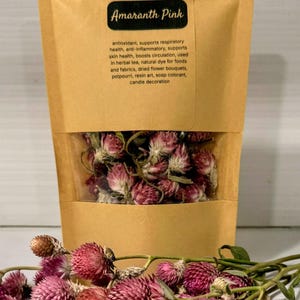 Pode incluir: Um saco de papel kraft com uma janela transparente cheio de flores de amaranto rosa secas. Flores de amaranto soltas estão espalhadas na frente. O saco tem o texto "Amaranth Pink" e "Dried Flowers".