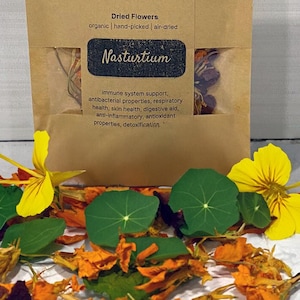 Puede incluir: Una pequeña bolsa de papel marrón con una etiqueta blanca que dice "VOMOV vomovplants.com Flores secas orgánicas | recogidas a mano | secadas al aire Capuchina apoyo del sistema inmunológico, propiedades antibacterianas, salud respiratoria, salud de la piel, ayuda digestiva, antiinflamatorio, antioxidante, propiedades desintoxicantes", con flores de capuchina secas que se caen de la bolsa sobre una superficie blanca.