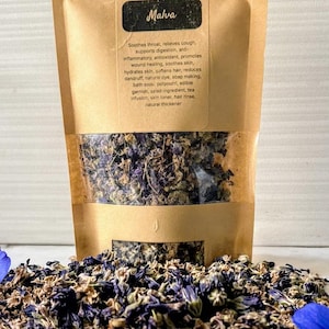 Può includere: Un sacchetto di carta marrone con fiori secchi "Malva" è posto sopra un mucchio degli stessi fiori, con due fiori blu su entrambi i lati. Il sacchetto ha una finestra trasparente. Lo sfondo è una superficie piastrellata bianca.