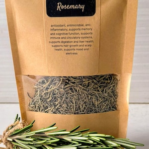 Puede incluir: Un paquete de hojas de romero secas con una ventana transparente, un manojo de romero fresco y un montón de romero seco. El paquete es marrón con la palabra "Rosemary" en una etiqueta negra. El texto del paquete dice "Dried Leaves".