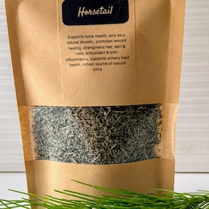 Puede incluir: Una bolsa de papel marrón con hojas secas de cola de caballo con una ventana transparente, un montón de hojas secas y una ramita de cola de caballo verde fresca. La bolsa tiene el texto "Dried Leaves" y "Horsetail".