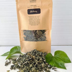 May include: A brown paper bag of dried mulberry leaves with a clear window, and a pile of loose leaves. The bag has the text "Mulberry" and information about the product. Green leaves are placed around the product.