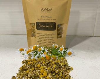 Könnte beinhalten: Ein beigefarbener Beutel mit getrockneten VoMoV-Kamillenblüten mit Sichtfenster. Das Etikett trägt die Aufschrift Dried Flowers, organic, hand-picked, air-dried und Chamomile. Ein Haufen loser, getrockneter gelber und weißer Kamillenblüten liegt davor.
