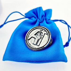 以下が含まれることがあります： 青いベルベットのポーチの上に置かれた、円形のデザインに「AT」の文字が刻まれた銀色のピン