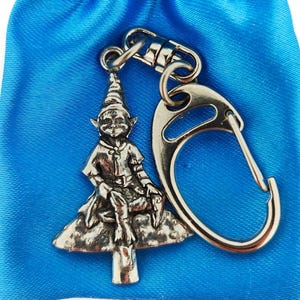 May include: A silver-toned keyring featuring a whimsical elf sitting on a tree stump. The elf is wearing a pointed hat and has a mischievous expression. The keyring has a silver-toned metal loop for attaching to keys.