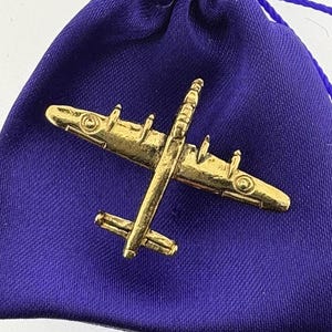 Pode incluir: Um alfinete de metal dourado de um avião bombardeiro vintage em uma bolsa de veludo roxo.